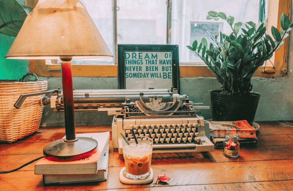 a typewriter, table lamp on two books, a glass of milk and a flower pot kept on a table for writing a blog